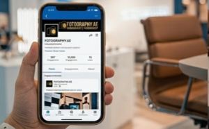 A hand holds a smartphone displaying the business profile page for "FOTOGRAPHY.AE." The screen shows engagement metrics and recent posts, set against a blurred background of a modern office or showroom.