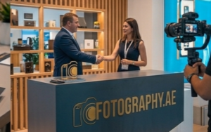 A close-up of a reception desk branded with "FOTOGRAPHY.AE." Behind the desk, a man in a suit and a woman in a dark dress shake hands, while a professional camera on a gimbal films them from the right.