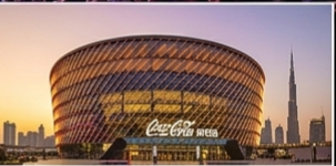  A vibrant sunset view of the Coca-Cola Arena in Dubai, showcasing its glowing amber facade with the Burj Khalifa and city skyline in the background.