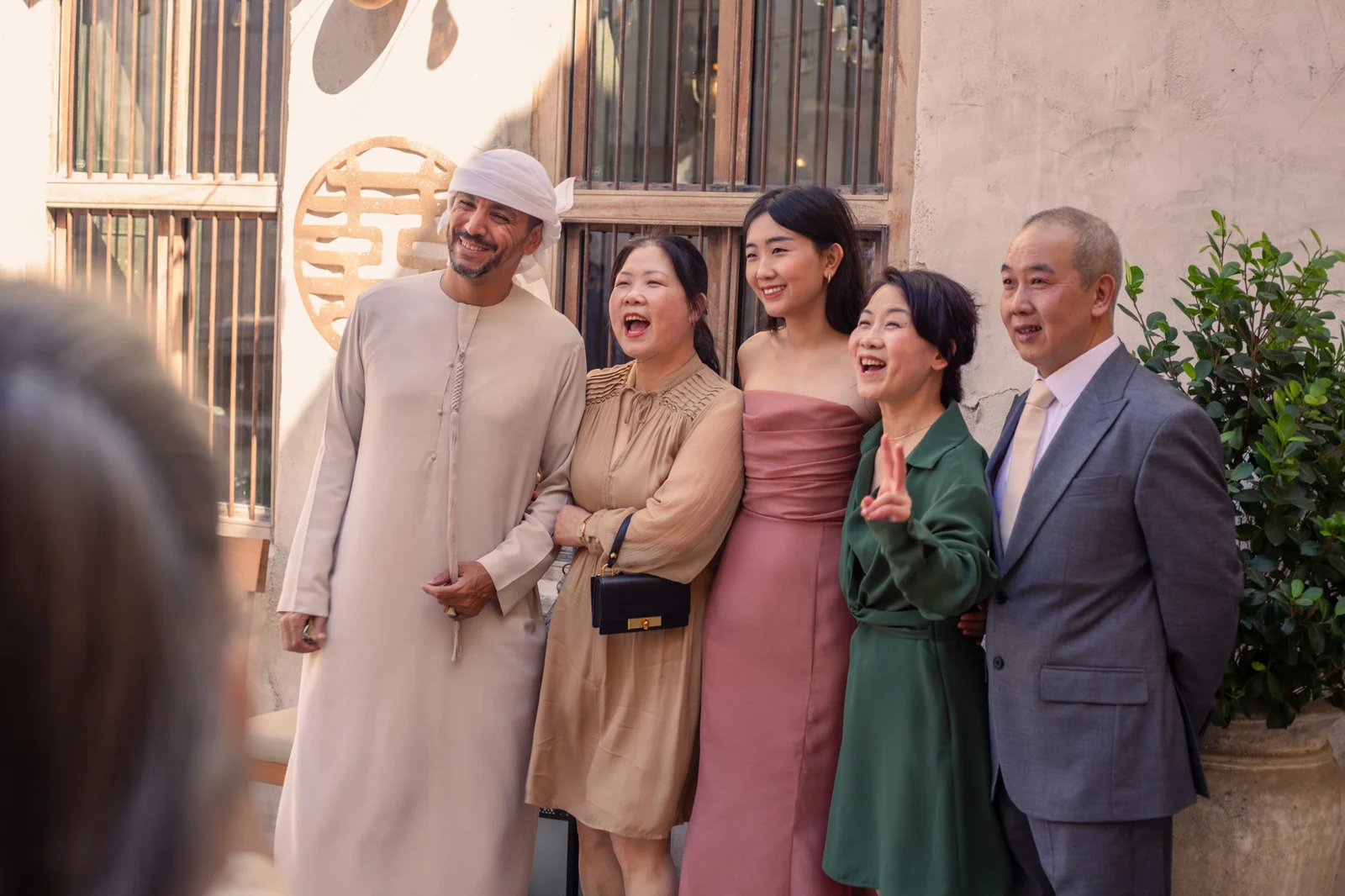 A diverse group of five people, including a man in traditional Emirati attire and four others in semi-formal Western-style clothing, posing and laughing together in front of a rustic building with a wooden-slatted window.