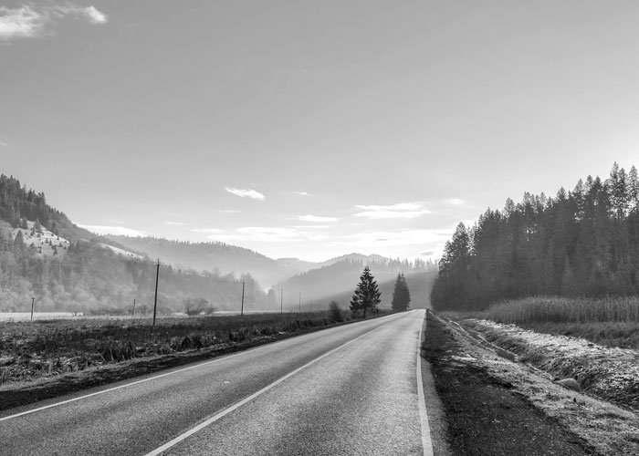 A monochrome photograph of a two-lane asphalt road stretching toward misty mountains. Dense evergreen forests line the right side of the road, while open fields and rolling hills sit to the left under a vast, clear sky.