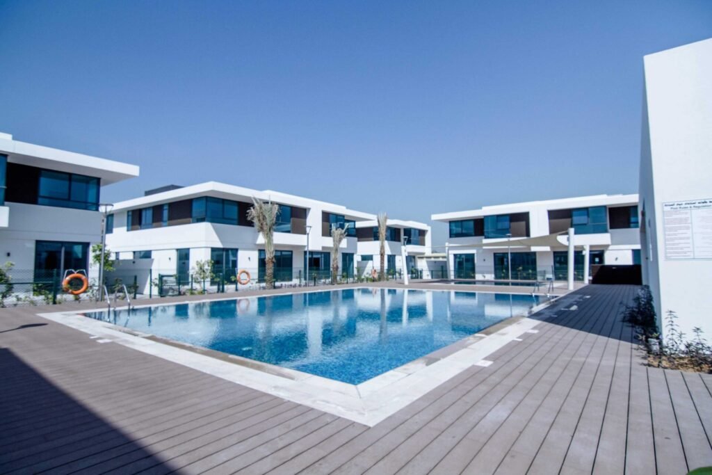 Modern luxury villas surrounding a large outdoor swimming pool with wooden decking under a clear blue sky