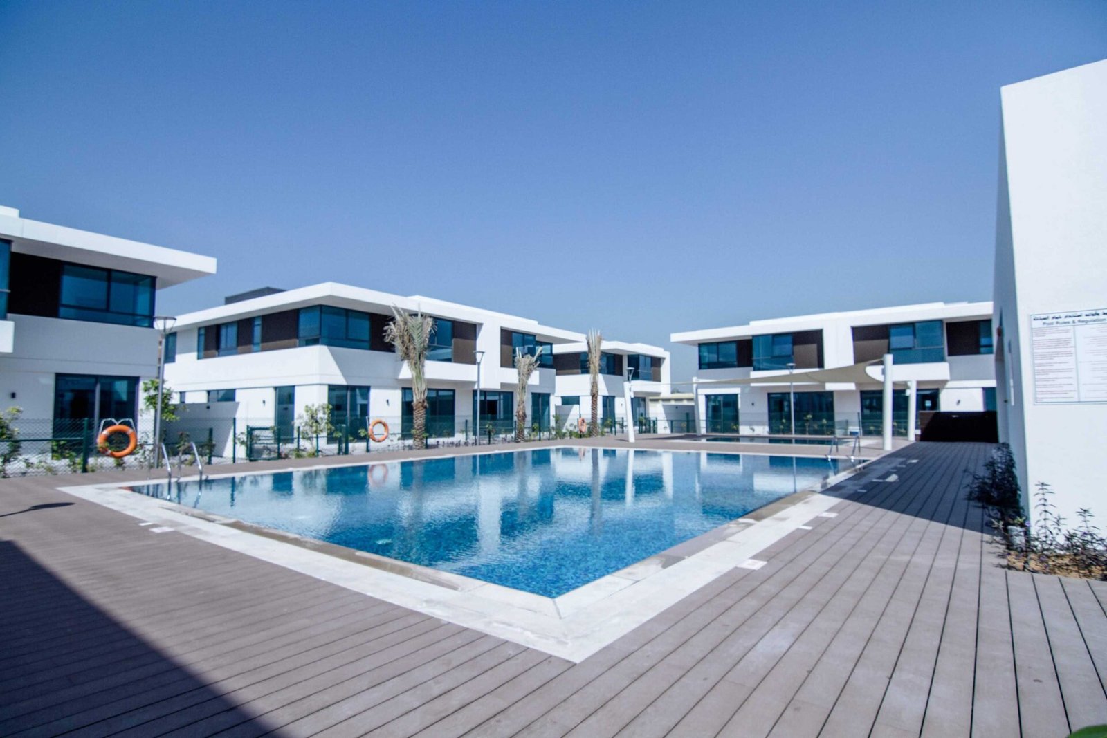 Modern luxury villas surrounding a large outdoor swimming pool with wooden decking under a clear blue sky