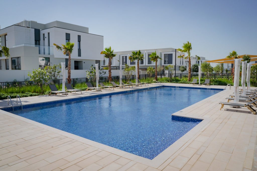 A large, rectangular blue-tiled swimming pool surrounded by a light-colored stone deck. The background features modern white multi-story residential buildings, palm trees, and white pool umbrellas under a clear, bright sky.