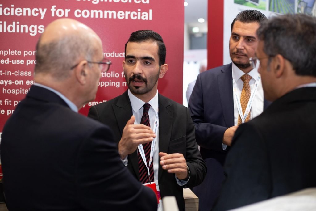 Three men in professional business suits engaged in a serious conversation at a trade show or exhibition. A red backdrop with white text regarding "efficiency for commercial buildings" is visible behind them.