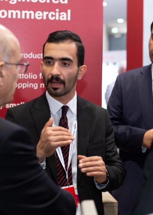 Three men in professional business suits engaged in a serious conversation at a trade show or exhibition. A red backdrop with white text regarding "efficiency for commercial buildings" is visible behind them.